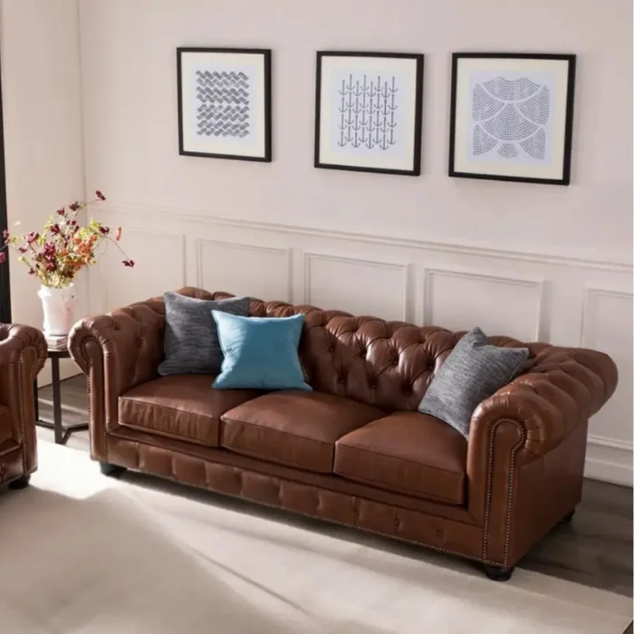 Windsor-Leatherite-Chesterfield-Sofa-details Brown Chesterfield sofa with tufted back, rolled arms, and nailhead trim, styled with gray and blue cushions beneath three framed artworks in a bright living room.